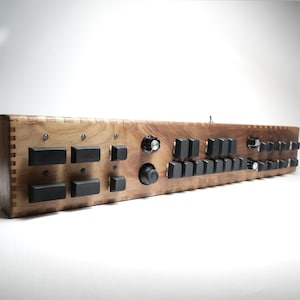 Op de afbeelding: Een lange, rechthoekige houten audiocontroller met een natuurlijke houtnerfafwerking en hoeken met vingerlassen. Het voorpaneel is uitgerust met meerdere zwarte rechthoekige knoppen, diverse draaiknoppen en een enkele tuimelschakelaar.