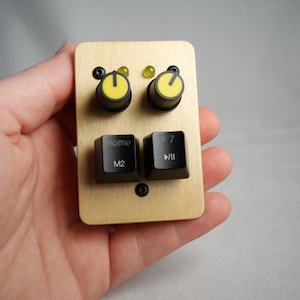 Puede incluir: Un pequeño dispositivo rectangular de madera con dos diales con perillas amarillas y dos teclas de teclado negras con las etiquetas "Home M2" y "F7 >/II". El dispositivo también tiene dos pequeñas luces verdes y se sostiene en una mano.