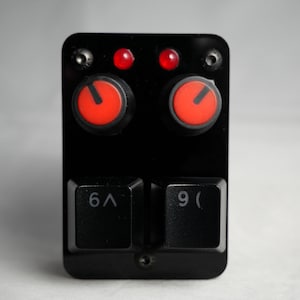 Puede incluir: Dispositivo electrónico negro con dos perillas rojas, dos luces rojas y dos teclas de teclado negras con los números "6" y "9". El dispositivo tiene forma rectangular.