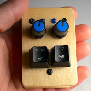 Puede incluir: Un dispositivo de madera personalizado con dos perillas azules y dos teclas de teclado negras con las etiquetas "Home" y "F7 >/II". El dispositivo tiene un acabado de veta de madera clara y pequeños tornillos negros.