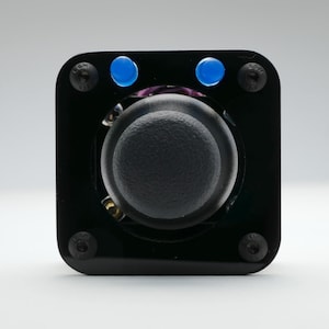 May include: A black square joystick controller featuring a central rounded black knob, two small blue circular buttons at the top, and four black screws securing the corners of the glossy black faceplate against a plain white background.