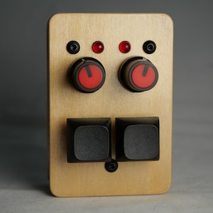 Puede incluir: Un dispositivo rectangular de madera con dos botones negros, dos diales con perilla roja y dos luces indicadoras rojas. El dispositivo tiene un acabado de veta de madera clara y tornillos negros. Los diales tienen un indicador rojo.