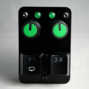Puede incluir: Dispositivo electrónico negro con botones de encendido y luces indicadoras verdes. Cuenta con dos teclas de teclado etiquetadas como "F1" y "3#". El dispositivo tiene forma rectangular con esquinas redondeadas y tornillos visibles.
