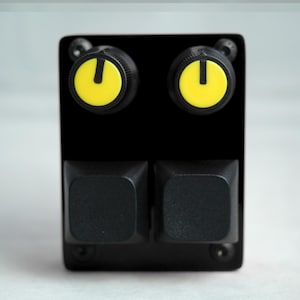 Puede incluir: Un dispositivo electrónico negro con dos perillas con detalles amarillos y dos botones cuadrados negros. Las perillas tienen un diseño de semicírculo amarillo. El dispositivo está montado sobre una base rectangular negra con tornillos en cada esquina.