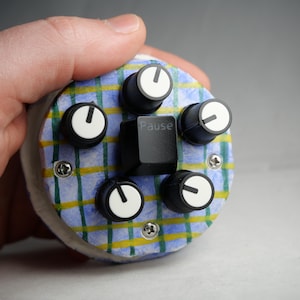 Puede incluir: Un dispositivo electrónico hecho a mano con un patrón de cuadros azules y amarillos. Cuenta con cinco perillas negras con indicadores blancos y una tecla negra con la etiqueta "Pause". El dispositivo está en la mano de una persona.