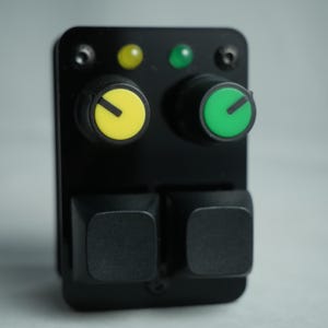 Puede incluir: Un dispositivo electrónico negro con dos perillas amarillas y verdes, dos botones cuadrados negros y dos luces indicadoras. Las perillas tienen una base negra con tapas amarillas y verdes. Los botones son cuadrados y negros.