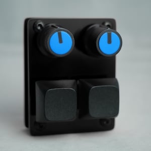 Puede incluir: Un panel de control negro con dos perillas con detalles azules y dos botones cuadrados negros. Las perillas tienen un exterior negro con un centro azul y una línea indicadora blanca. Los botones son cuadrados y negros.