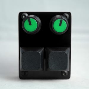 Puede incluir: Un dispositivo electrónico negro con dos perillas verdes y dos botones cuadrados negros. Las perillas tienen un indicador verde. El dispositivo está montado en una placa negra con tornillos.