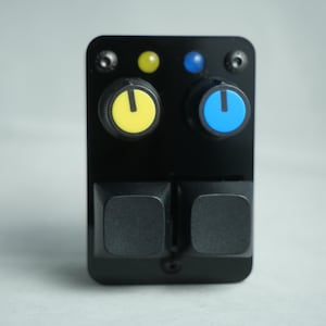 Puede incluir: Un panel de control electrónico negro con dos perillas amarillas y azules, dos botones cuadrados negros y dos tornillos pequeños. El panel también tiene dos pequeñas luces indicadoras, una amarilla y otra azul.