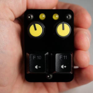 Puede incluir: Un panel de control negro con dos perillas amarillas y dos luces indicadoras amarillas. El panel también presenta dos teclas de teclado etiquetadas como "F10" y "F11" con símbolos de volumen. El dispositivo se sostiene en una mano.