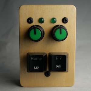 Puede incluir: Un dispositivo rectangular de madera con dos perillas verdes, dos luces verdes y dos teclas de teclado negras etiquetadas "Home" y "F7". El dispositivo tiene un acabado de veta de madera clara y tornillos negros.