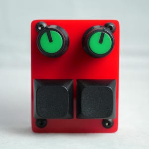 Puede incluir: Un dispositivo electrónico rojo con dos botones cuadrados negros y dos perillas verdes. Las perillas están en la parte superior y los botones en la parte inferior. El dispositivo está montado con cuatro tornillos negros.