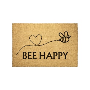 May include: A brown coir doormat with the text "BEE HAPPY" in black lettering. A black and white illustration of a bee flying in a dotted line path with a heart at the end of the path.