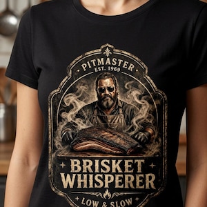 May include: A black t-shirt featuring a graphic print of a bearded person wearing sunglasses and an apron, standing behind a piece of brisket. The text reads PITMASTER EST. 1969, BRISKET WHISPERER, and LOW & SLOW, surrounded by swirling smoke.