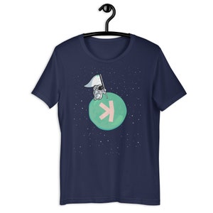 May include: A navy blue t-shirt featuring a cartoon astronaut standing on a green planet with a pink arrow. The astronaut is holding a white flag. The background is a dark blue space with white stars.