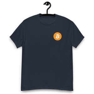 Pode incluir: Uma camiseta azul escura com um pequeno círculo laranja contendo um símbolo Bitcoin branco.