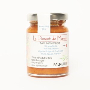 Pode incluir: Um frasco de vidro de molho picante de cor laranja com um rótulo branco que diz "Le Piment de Maman" e "Sans Conservateurs". O rótulo também lista os ingredientes: "Piment Antillais", "Oignon Rouge de Toulouges" e "Huile Rouge de Bénin". O frasco é rotulado como "FORT" e tem um logotipo de palmeira.