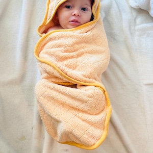 May include: A baby wrapped in a peach-colored hooded towel with yellow trim. The baby is swaddled in the towel, with only the face visible. The towel is soft and fluffy, and the baby is looking at the camera.
