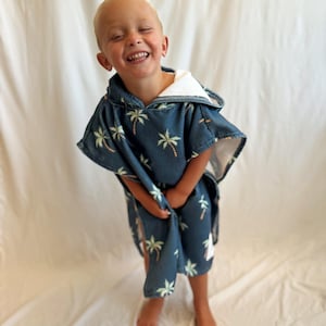 May include: A navy blue hooded beach towel with a palm tree pattern. The towel is worn by a child. The hood is lined with white terry cloth. The child is smiling and appears to be happy.