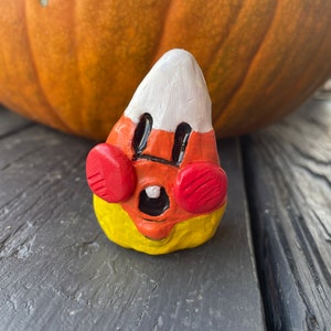 May include: A hand-painted candy corn figurine with a white top, orange middle, and yellow bottom. The figurine has a cute face with red cheeks and black eyes.