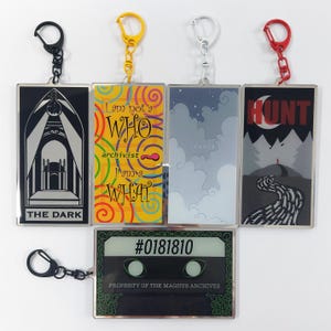 May include: A collection of rectangular keychains with various designs and text. The keychains feature images and phrases such as "THE DARK", "I am not a WHO, archivist, I am a WHAT", "lonely", "HUNT", and a cassette tape design with "#0181810". Each keychain has a different colored clasp.