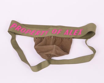 May include: Olive green jockstrap with adjustable straps and the text "PROPERTY OF ALEX" in pink lettering.