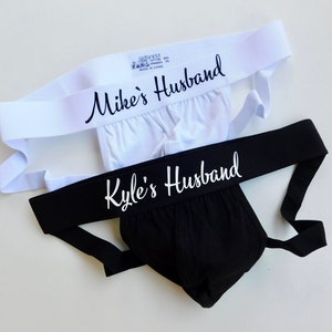 May include: Two jockstraps with white elastic bands. The top strap has the text "Mike's Husband" in black script. The bottom strap has the text "Kyle's Husband" in white script.