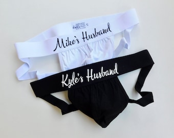 May include: Two jockstraps with white elastic bands. The top strap has the text "Mike's Husband" in black script. The bottom strap has the text "Kyle's Husband" in white script.