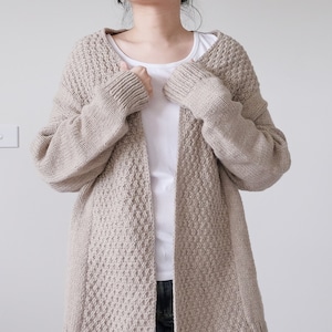 Könnte beinhalten: Ein beigefarbener Strickcardigan mit einem strukturierten Muster. Der Cardigan ist vorne offen und hat lange Ärmel.
