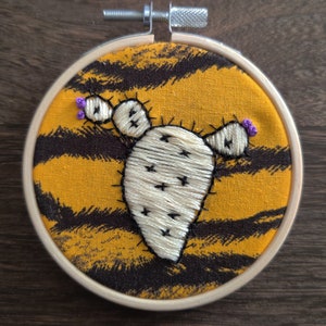May include: Embroidered cactus with purple flowers on a yellow and black striped fabric in a wooden embroidery hoop.