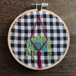 May include: A green plant in a teal pot hangs from a pink macrame hanger on a black and white checkered background.