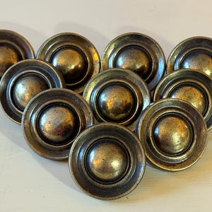 May include: A collection of ten antique brass cabinet knobs. Each knob has a circular design with a raised center and a ridged outer edge. The knobs have a tarnished, aged appearance, suggesting vintage hardware.
