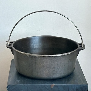 May include: A round, dark gray cast iron pot with a smooth interior and a thick, curved metal bail handle attached to two side lugs. The pot features a wide, open top and a flat base, presenting a heavy-duty construction with a matte metallic finish.