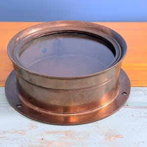 May include: A circular nautical porthole window featuring a polished copper frame with a clear glass center. The metal frame includes a wide, flat mounting flange with pre-drilled holes around the perimeter for secure installation on a wall.