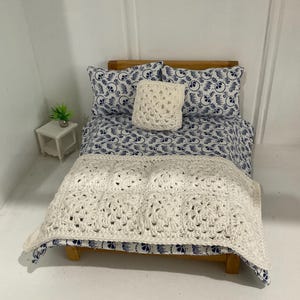 May include: A miniature wooden bed with a white crocheted blanket and a blue and white patterned bedding set. The bed has a small white crocheted pillow and a white bedside table with a small green plant.