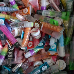 Peut inclure: Un assortiment de tubes et pots de gloss de différentes couleurs, dont rose, orange, vert et transparent. Certains tubes ont des étiquettes avec du texte, et les produits semblent être des articles cosmétiques.
