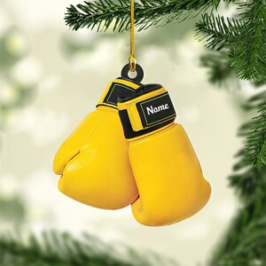 May include: Yellow boxing gloves ornament with black accents and the word "Name" on the wrist. The gloves are hanging from a gold string, perfect for a Christmas tree or sports-themed decor.