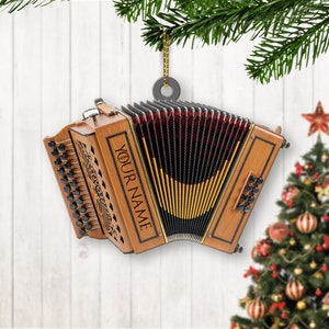 May include: A wooden accordion Christmas ornament with the text "YOUR NAME" engraved on the side. The ornament has black and gold bellows and is hanging from a gold string. A Christmas tree is in the background.