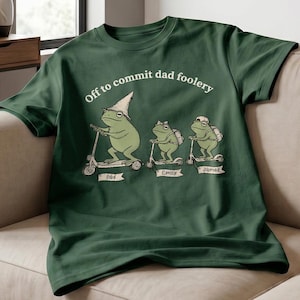 May include: Dark green t-shirt with the phrase "Off to commit dad foolery" above a graphic of three frogs on scooters. The frogs are labeled "Dad", "Emily", and "James".