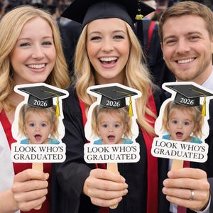 May include: Three people smiling and holding graduation photo props. Each prop features a baby's face wearing a graduation cap with "2026" and the text "LOOK WHO'S GRADUATED" on a white background.