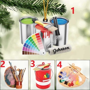 May include: A set of artist-themed ornaments. The ornaments include paint cans with green, red, and blue paint, a color palette, a paint roller with the name "Johnson", and paintbrushes. The ornaments are hanging from a Christmas tree.