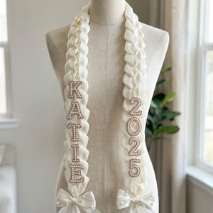May include: Cream-colored graduation sash with the name "KATIE" spelled out in rhinestone letters and the year "2025" in rhinestone numbers. The sash is adorned with white bows and pearls.