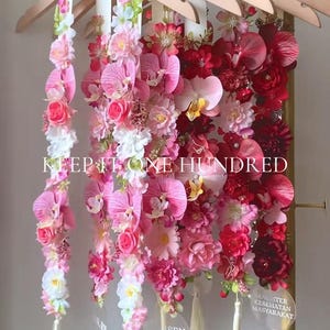 May include: Wooden hangers adorned with floral arrangements in shades of pink and red. The hangers are suspended from a gold-colored rod. The text "BLOOM" and "KEEP IT ONE HUNDRED" are visible.