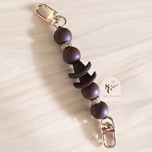 May include: A brown and gold charm necklace with a hat charm and rhinestones. The necklace is made of silicone beads and has a gold lobster clasp.