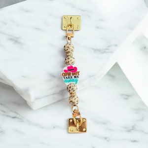 May include: A gold-colored charm with a rectangular clasp at each end, connected by a chain of leopard-print beads. A charm with a pink cowboy hat and the text "Cowboy Take Me Away" is in the center.