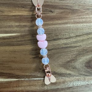 May include: A rose gold pacifier clip with heart-shaped accents. The clip features a series of pink heart-shaped beads and blue speckled beads, separated by a row of clear beads. The clip is designed to attach to a pacifier.
