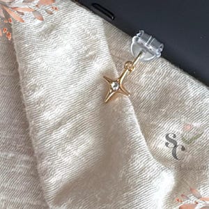 May include: A gold-colored star-shaped earring with a clear gem in the center. The earring is attached to a clear plastic backing. The earring is displayed on a white, textured fabric.