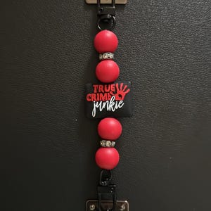 May include: A black and red keychain with the text "TRUE CRIME junkie". The keychain features red beads, silver accents, and a black rectangular charm with a red handprint. The keychain is attached to a silver metal clasp.