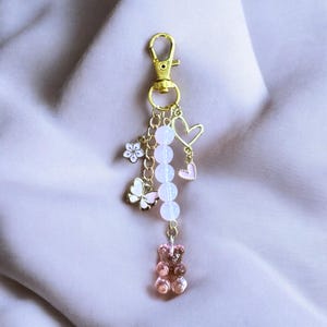 May include: A gold-colored keychain with a lobster clasp, adorned with charms. Charms include a flower, butterfly, heart, and a gummy bear. The keychain features a chain with white beads and pink accents, set against a soft pink background.