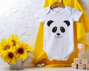 May include: White baby onesie with a black and white panda face graphic. The onesie is hanging on a wooden hanger against a white textured wall. A bouquet of yellow sunflowers is in a white watering can in the foreground.